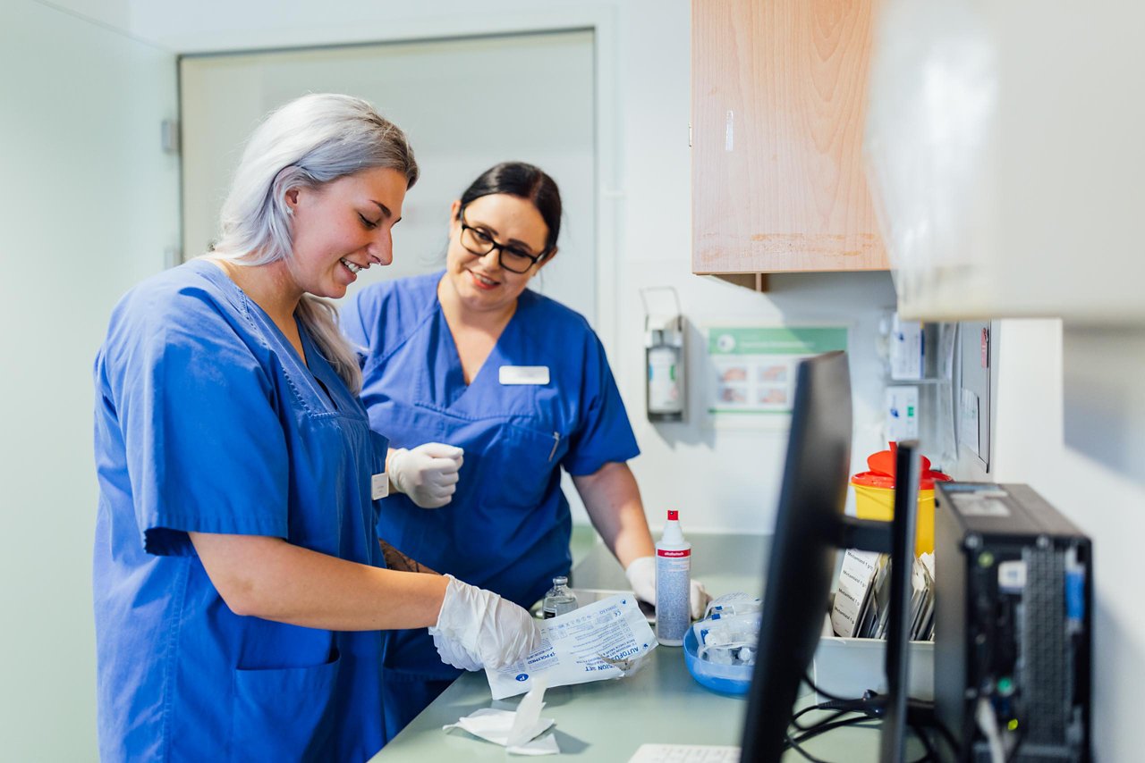 Ein Krankenpflegehelfer schaut sich unter Anleitung einer Praxisanleiterin die Vitalzeichen eines Patienten an.