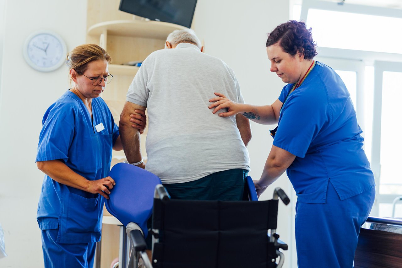 Claudia und Natalie helfen einem Patienten dabei, sich in den Rollstuhl zu setzen