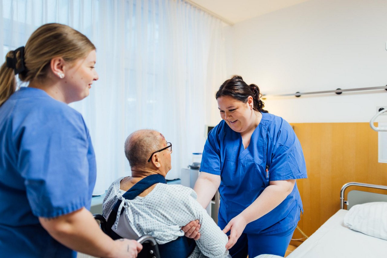 Eine KPH Auszubildende hilft einem Patienten dabei, vom Rollstuhl ins Bett zu kommen.