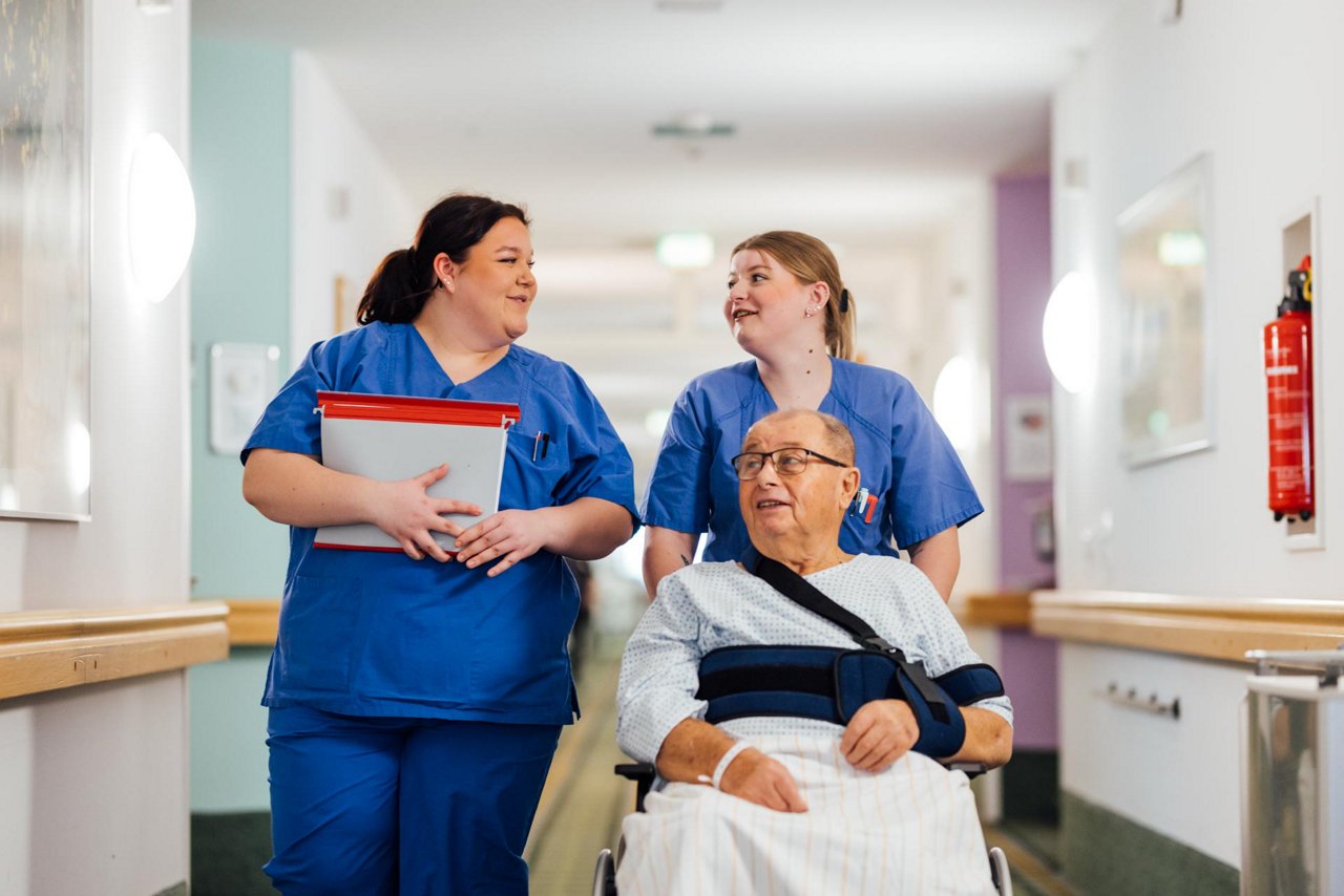 Zwei KPH Azubis transportieren einen Patienten mit einem Rollstuhl