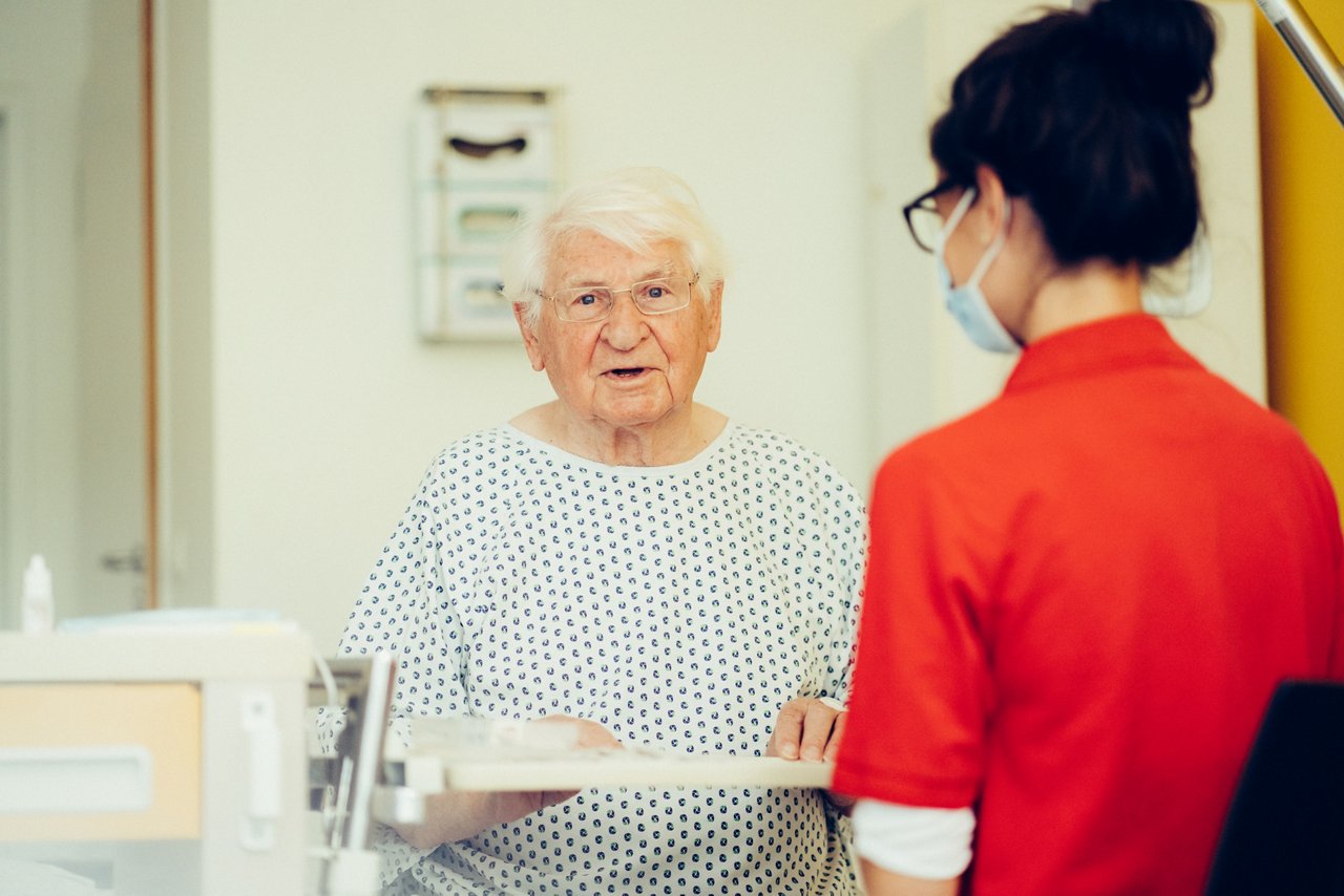 Logopädin und Patient sitzen am Krankenbett, der Patient blickt in die Kamera