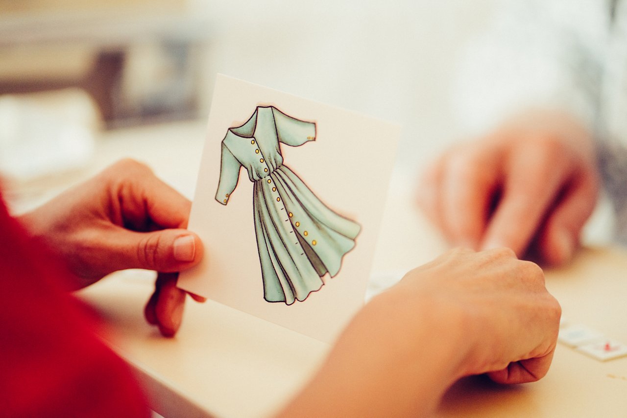 Es wird eine Karte hochgehalten auf dem ein Kleid gezeichnet ist, mehrere Hände zeigen auf Buchstaben auf dem Tisch