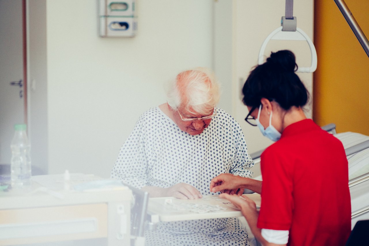 Patient und Logopädin sitzen am Krankenbett und sortieren Buchstaben auf kleinen Kärtchen