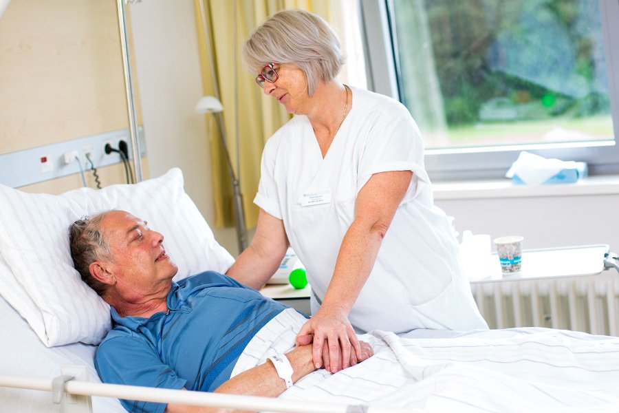 Schwester steht am Patientenbett und hält die Hände 