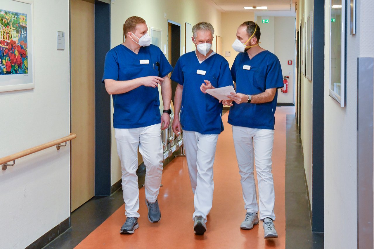 Drei Ärzte der Unfallchirurgie mit FFP2-Maske gehen über den Flur und besprechen Dokument in der Hand