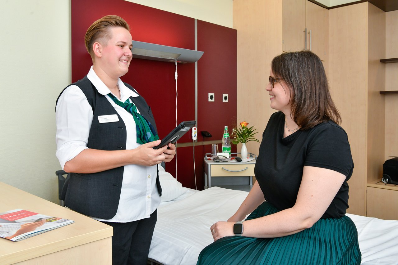 Servicekraft befragt Patientin im Patientenzimmer mit Servicemonitor am Tablet nach Zufriedenheit