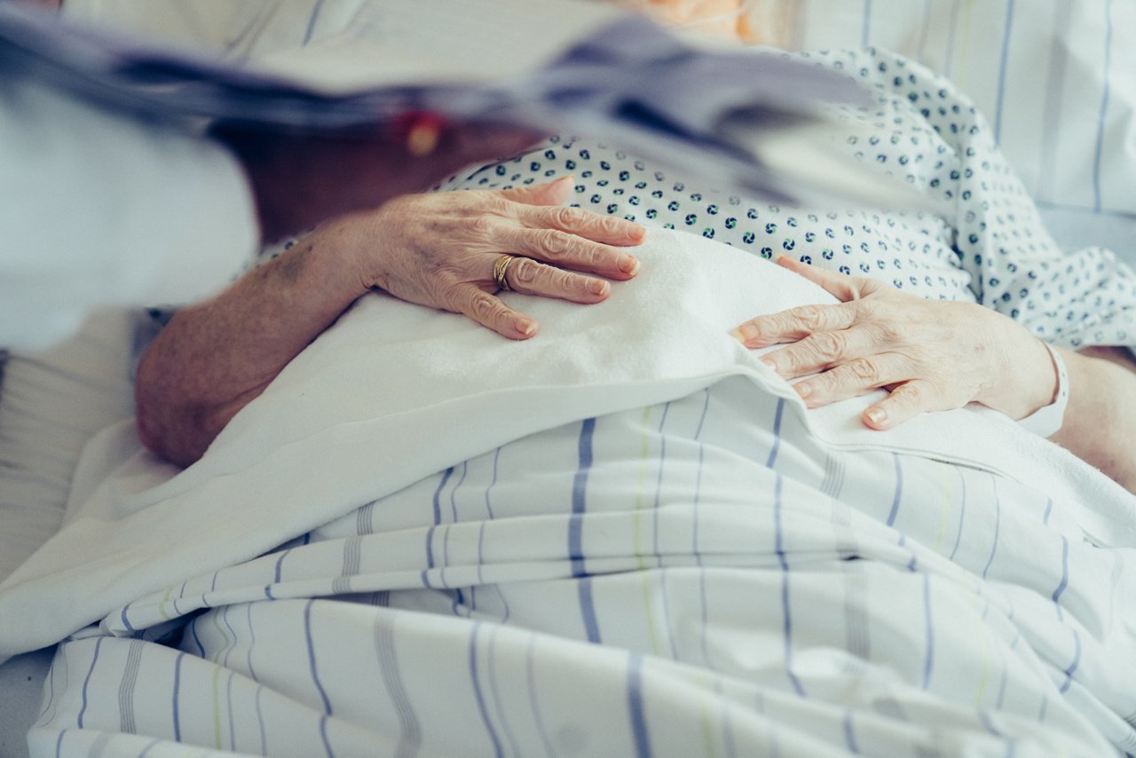 Hände von Patient:in im Krankenbett liegen auf dem Bauch, Hand mit Unterlagen im Vordergrund 