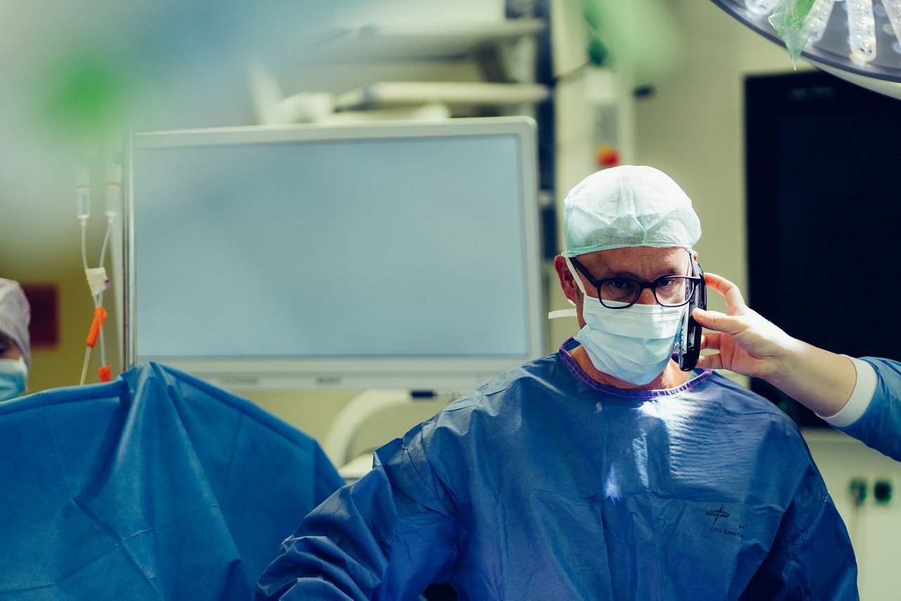 Arzt in OP Kleidung bekommt Telefon von Assistenten an das Ohr gehalten