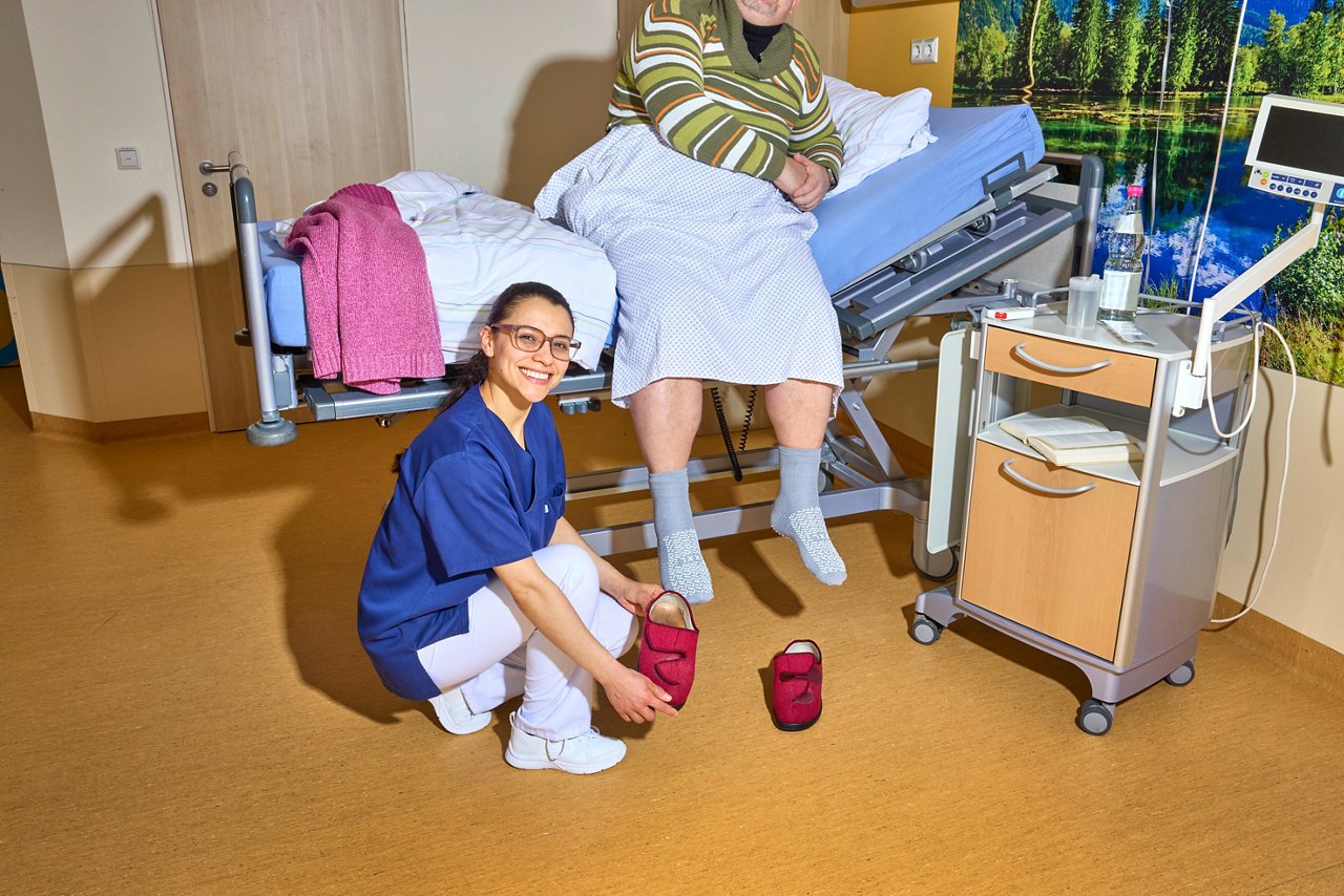 Eine freundliche Pflegekraft hilft einem Patienten im Krankenhaus, indem sie ihm Hausschuhe reicht. Der Patient sitzt auf einem verstellbaren Bett, umgeben von einer beruhigenden Umgebung.