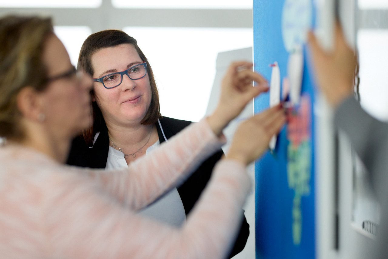 Kursleiterin beobachtet Seminarteilnehmerin die einen Zettel an eine Pinwand hängt