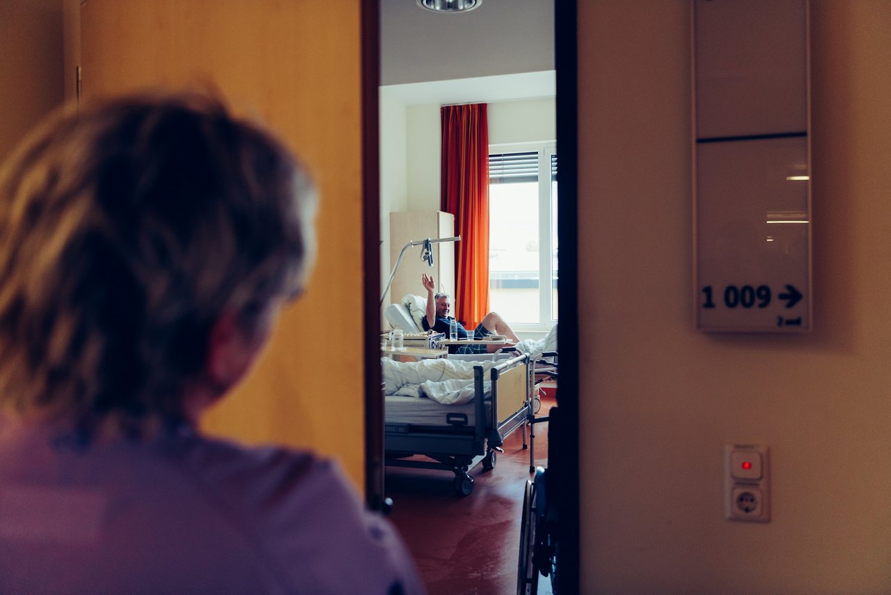 Patient im Krankenbett winkt der Reinigungskraft auf dem Flur