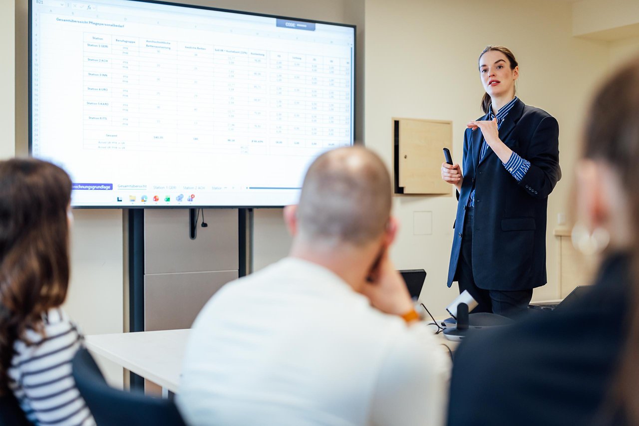 Eine Geschäftsfrau präsentiert in einem modernen Konferenzraum Daten auf einem Bildschirm. Die Zuhörer sind aufmerksam und interagieren mit der Präsentation.