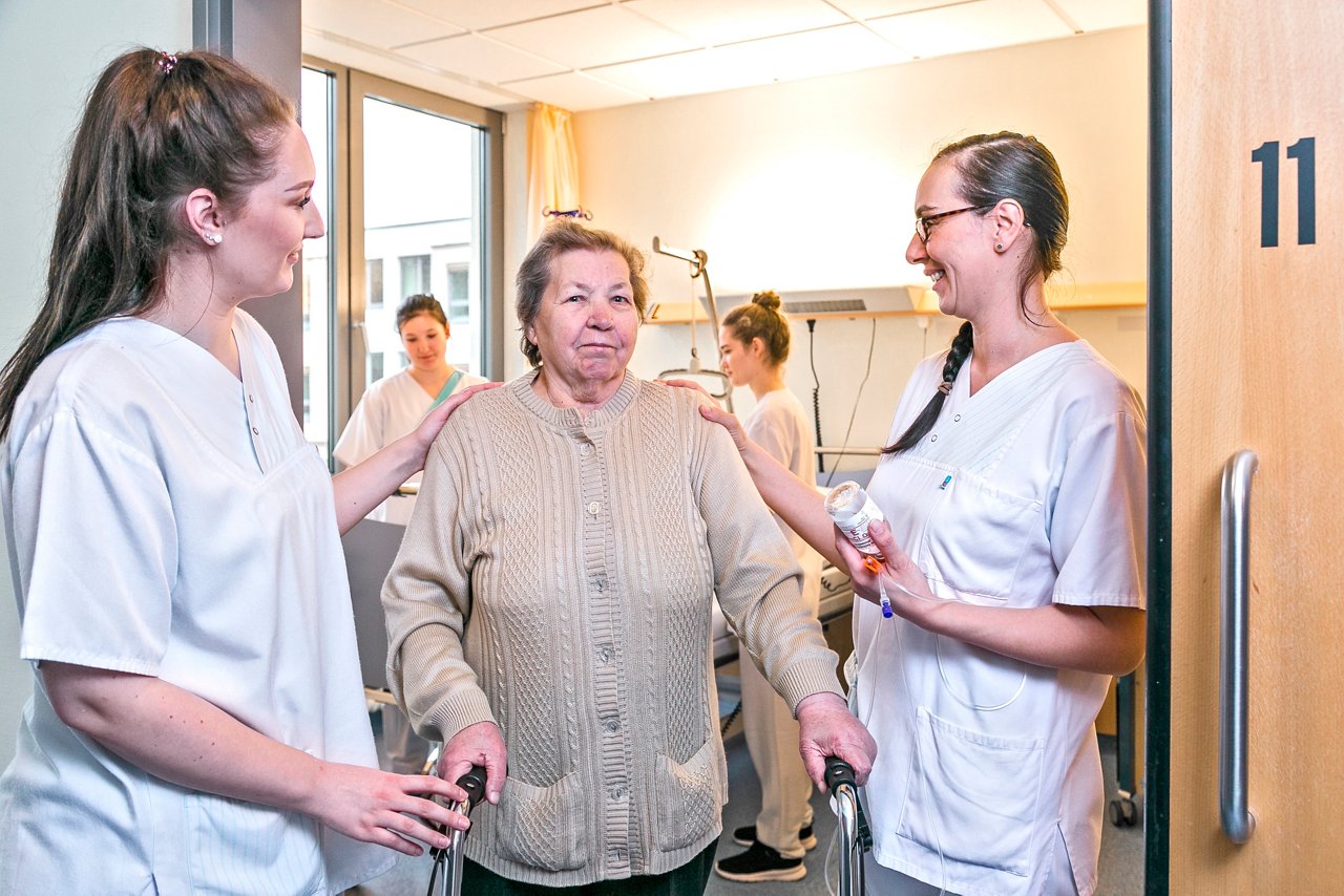 Auszubildende der Pflege auf der Schulstation helfen Patientin mit Rollator