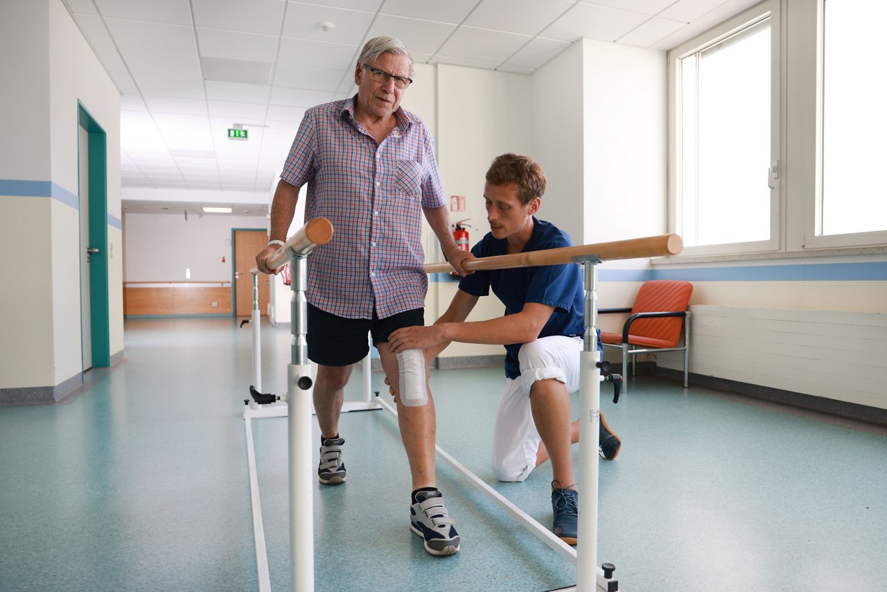 Physiotherapeut trainiert mit Patienten am Barren, und korrigiert die  Stellung seines linken Beines