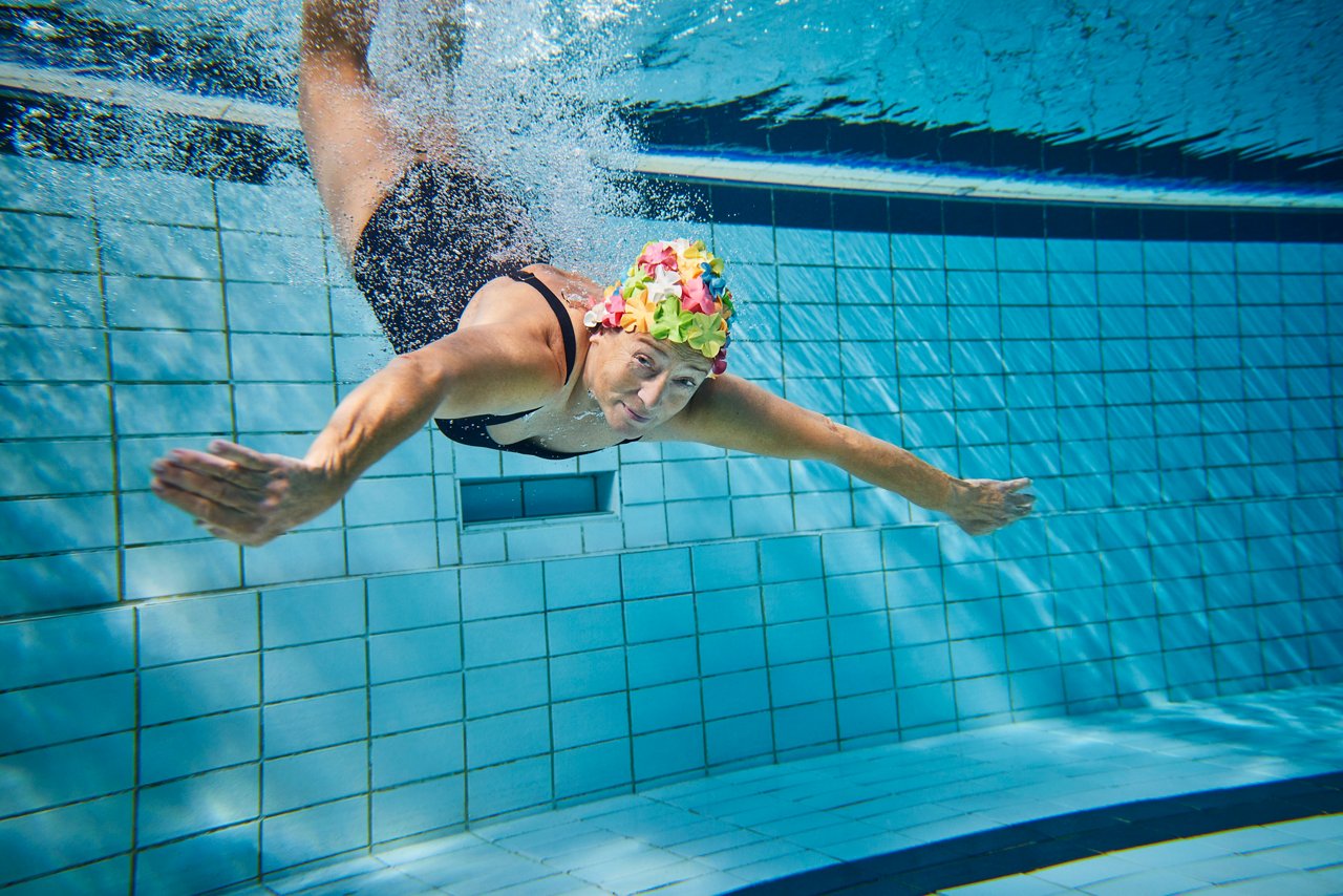 Frau mit bunter Badekappe taucht im Schwimmbad Patientenkampagne 2022