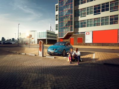 Hyundai KONA Electric stoi zaparkowany przy ładowarce, a obok na chodniku siedzi para ludzi.