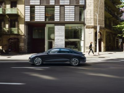 The all-electric Hyundai IONIQ 6 driving on a city street past a modern building.