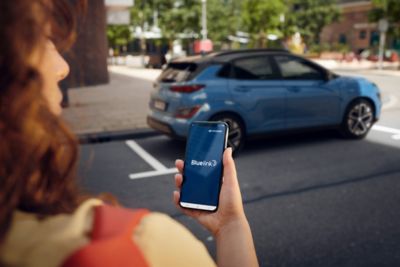 Eine Frau hält ein Smartphone mit Bluelink Logo in der Hand, im Hintergrund steht ein Hyundai KONA Elektro.