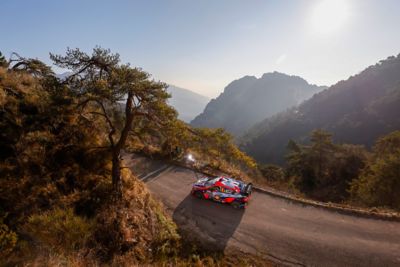 Hyundai Motorsport driver Oliver Solberg's i20 N WRC Rally1 driving on curvy road in the mountains.