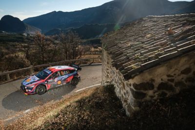 The Hyundai i30 N WRC rally race car driving around a corner next to an old building.