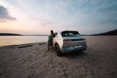 The Hyundai IONIQ 5 electric midsize CUV parked at the beach with a woman leaning against it, enjoying the sundown.