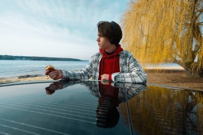 A young person, leaning on the solar roof of the Hyundai IONIQ 5 midsize CUV, parked at a beach.