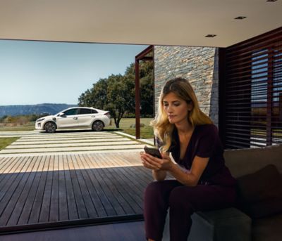 Mladá žena sledující svůj chytrý telefon za velkým oknem s venku zaparkovaným novým vozem Ioniq