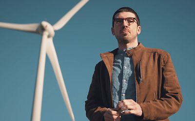 En mann står foran en vindturbin som produserer fornybar energi til IONITY-stasjonene. Foto.