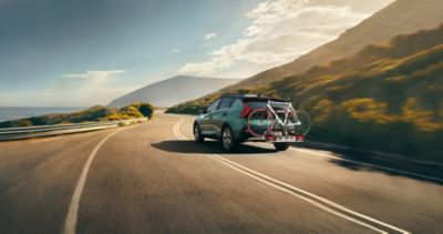 The Hyundai BAYON with the accessories tow bar and bike carrier driving on a country road.