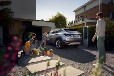 The Hyundai TUCSON Plug-in Hybrid parked next to its home charging box and a family playing.