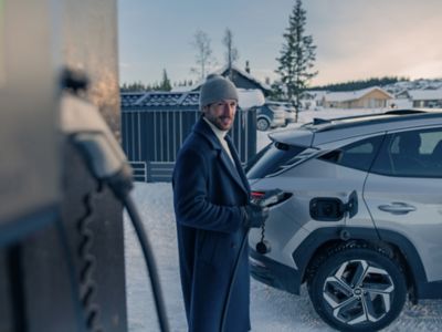 Ein Mann steht im Winter in einer Einfahrt, bereit einen Ladestecker in seinen Hyundai zu stecken.