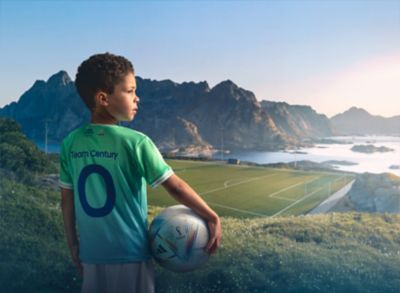 Garçon vêtu du maillot Goal of the Century Hyundai et tenant un ballon de foot, devant un paysage montagneux.