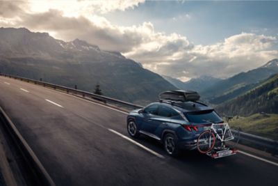 Hyundai Tucson with the accessories tow bar, bike carrier and roof box on a country road.