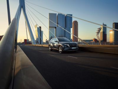 The Hyundai TUCSON Hybrid driving over a bridge in the city with the skyline in the background.