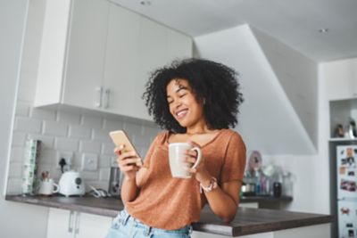 Woman smiling in a kitchen, drinking coffee and checking the smartphone. 