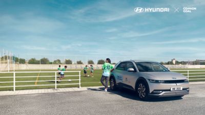 Nadia Nadim membro di Team Century di Hyundai osserva i bambini che giocano a calcio appoggiata a Hyundai IONIQ 5