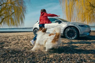 Hyundai IONIQ 5 crossover 100% elettrico parcheggiato accanto ad  un lago con una ragazza che gioca con un cane