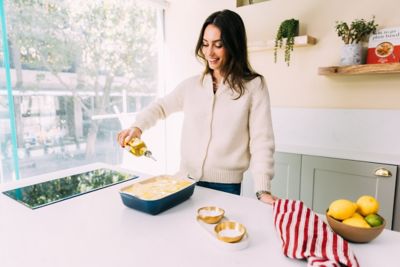 Ella Mills dans sa cuisine, face à une grande fenêtre. Elle verse un filet d’huile d’olive sur un plat prêt à être enfourné. Elle porte un cardigan blanc et un jean bleu.