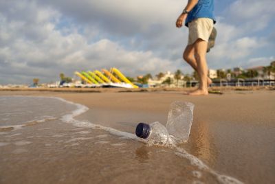 Persona camina por la orilla de una playa con una botella de plástico abandonada en primer plano.