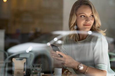 Una mujer mira a su Hyundai desde la ventana de una cafetería mientras sostiene su teléfono.