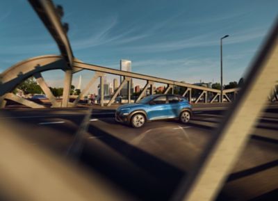 Nuevo Hyundai KONA en color Surfy Blue desde el lateral conduciendo sobre un puente.