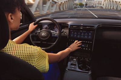 Eine Frau am Steuer eines Hyundai TUCSON greift an eine Taste der MIttelkonsole während sie über eine Brücke fährt.