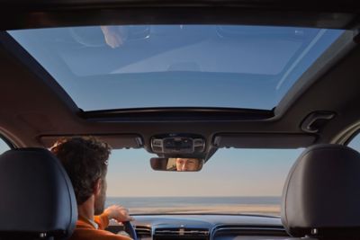 Blick durch die Frontscheibe und das Panorama-Glasdach eines Hyundai TUCSON. Ein Mann sitzt am Steuer.