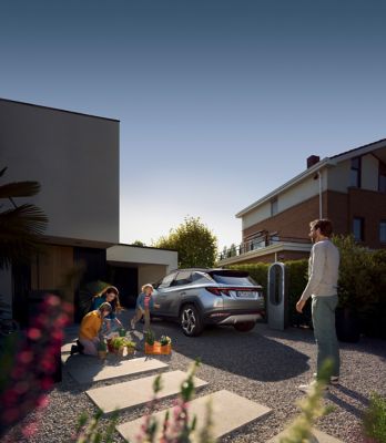 The Hyundai TUCSON Plug-in Hybrid parked next to its home charging box and a family playing.