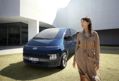 Eine junge Frau steht neben einem Hyundai STARIA vor einem modernen Gebäude. 