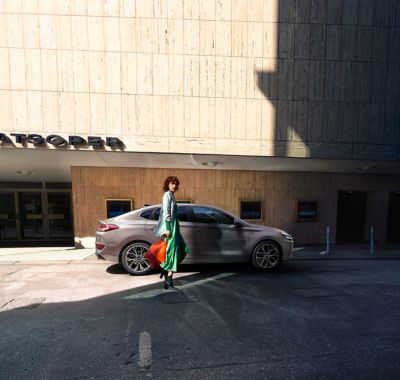 Una mujer moderna con vestido verde y una bolsa de compras roja camina hacia el Hyundai i30 Fastback.