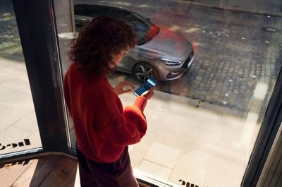 Mujer junto a una ventana, mirando la aplicación Bluelink en su teléfono, con el Hyundai i30 Fastback aparcado en la calle.