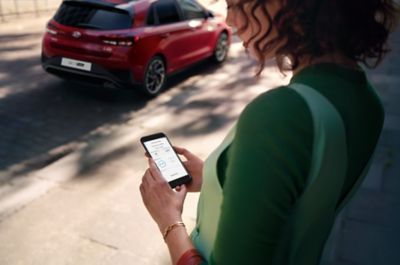 A woman looking at the Hyundai Bluelink App on her smartphone, her car is parked outside.