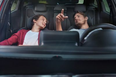 Father and son sitting in the front of a parked Hyundai i30 Fastback. Their seats are laid back and they are talking to each other.