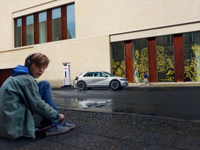 Ein Junge mit Kopfhörer vor einem öffentlichen Gebäude. Ein IONIQ 5 lädt an einer IONITY Ladesäule.